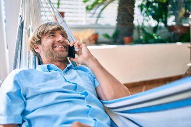Orta yaşlı yakışıklı bir adam evinin terasında, bir hamakta uzanıp telefonda konuşuyor.