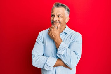 Orta yaşlı, gri saçlı, sıradan kıyafetler giyen, elleri çenesinde soru soran, düşünceli bir adam. Düşünceli bir yüzle gülümsüyor. şüphe kavramı. 