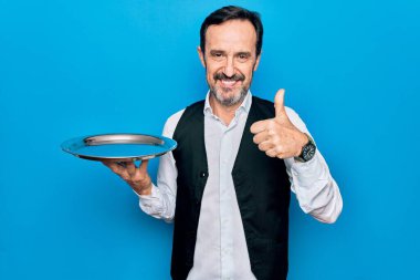 Orta yaşlı, yakışıklı garson, mavi arka planda duran tepsiyi tutuyor mutlu ve pozitif gülümsüyor, başparmağıyla mükemmel ve onay işareti yapıyor.