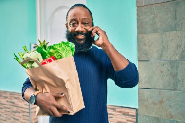 Elinde süpermarketten alınmış kağıt torba sakallı Afrikalı Amerikalı adam telefonda konuşuyor.