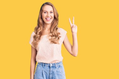 Sarışın, genç, beyaz, güzel bir kadın. Günlük kıyafetler giyiyor. İki numarayı gösteriyor ve gülümsüyor.. 