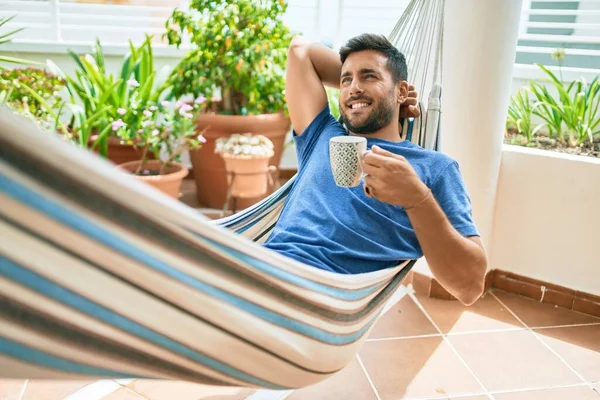 年轻的惊慌失措的男人在阳台上的吊床上放松地喝着一大杯咖啡 — 图库照片