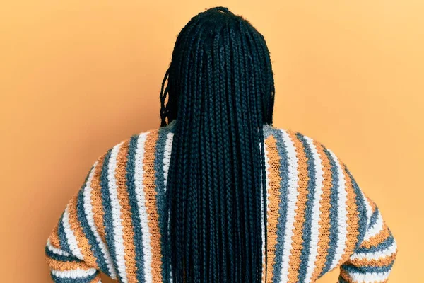 Saçları örgülü genç siyahi bir kadın sıradan bir kazak giyiyor. Arkaya bakıyor ve kollarını vücuduna dikiyor. 