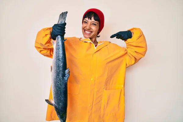 Beautiful brunettte fisher woman wearing raincoat holding fresh salmon pointing finger to one self smiling happy and proud