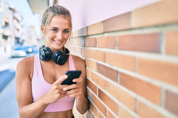 在城市街道上 年轻的金发女用智能手机和耳机靠在墙上 — 图库照片