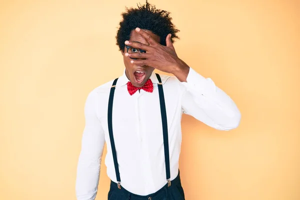 Handsome african american man with afro hair wearing hipster elegant look peeking in shock covering face and eyes with hand, looking through fingers with embarrassed expression.