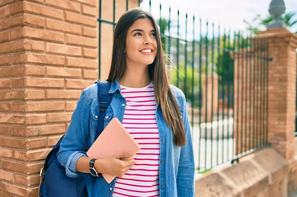 Young hispanic student girl smiling happy holding book at the city.