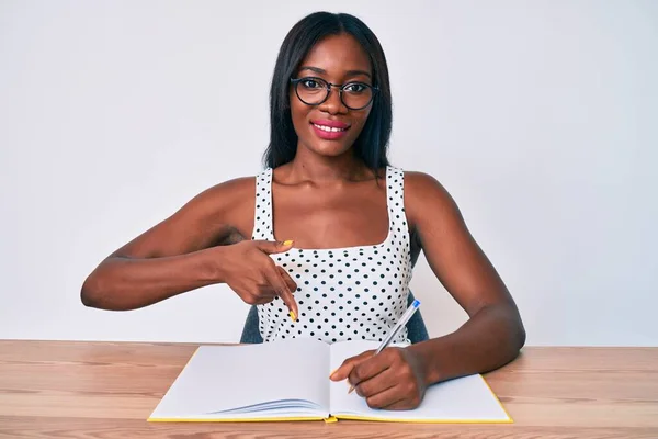 Young african american woman writing book sitting on the table smiling happy pointing with hand and finger