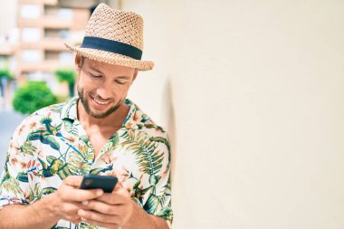 Beyaz tenli yakışıklı adam dışarıda mutlu bir şekilde gülümsüyor. Duvara yaslanmış akıllı telefon kullanıyor.