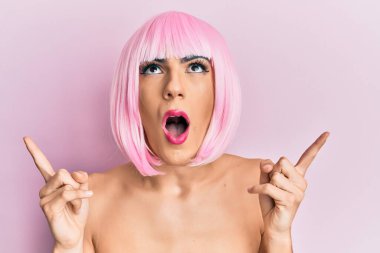 Pembe peruk takan genç adam şaşırmış ve şaşırmış bir şekilde yukarı bakıyor ve parmaklarıyla işaret ediyor ve kollarını kaldırıyor.. 