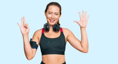 Güzel sarışın kadın spor kıyafetleri giyiyor ve kulaklık takıyor. Sekiz numaralı parmaklarını gösterip işaret ediyor. Kendinden emin ve mutlu bir şekilde gülümsüyor.. 