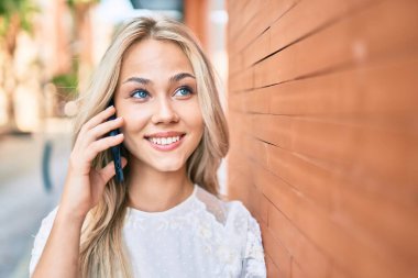 Kafkasyalı genç kız gülümsüyor. Duvara yaslanmış akıllı telefondan konuşuyor..