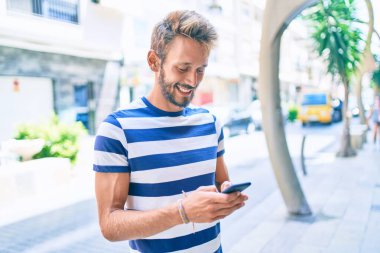 Sakallı yakışıklı beyaz adam dışarıda mutlu bir şekilde gülümsüyor. Akıllı telefon kullanıyor.