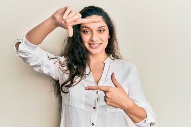 Ortadoğulu güzel bir kadın günlük kıyafetler giyip gülümsüyor ve elleri ve parmakları gülen yüzlü bir çerçeve yapıyor. yaratıcılık ve fotoğrafçılık kavramı. 