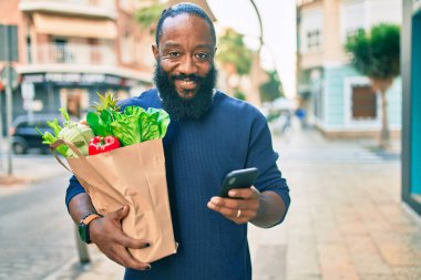 Sakallı Afrikalı Amerikalı adam elinde süpermarketten alınmış kağıt torbalarla akıllı telefon kullanıyor.