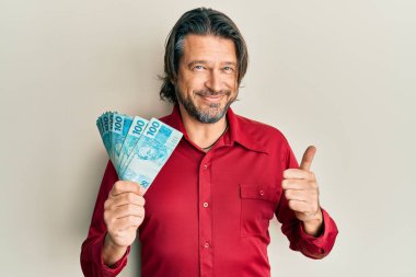 Orta yaşlı yakışıklı adam elinde 100 Brezilyalı gerçek banknot tutuyor mutlu ve pozitif gülümsüyor, başparmağıyla mükemmel ve onay işareti yapıyor. 