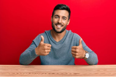 Masanın üzerinde sıradan giyinmiş yakışıklı İspanyol bir adam. Başarı işareti yapıyor. Elleriyle olumlu jest yapıyor, gülümsüyor ve mutlu. Neşeli ifade ve kazanan jest. 