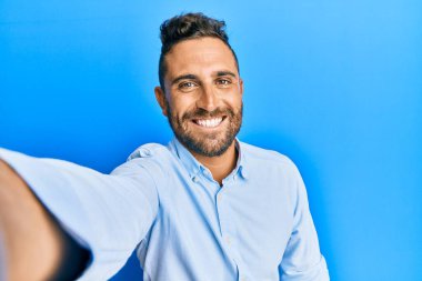 Sakallı yakışıklı bir adam akıllı telefonuyla fotoğraf çekiyor. Pozitif görünüyor ve mutlu görünüyor. Kendine güvenen bir gülümsemeyle dişlerini gösteriyor. 