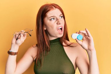 Young red head girl holding glasses and contact lenses angry and mad screaming frustrated and furious, shouting with anger. rage and aggressive concept. 