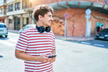 Beyaz adam mutlu bir şekilde gülümsüyor. Kulaklık takıyor, akıllı telefon kullanıyor ve şehirde yürüyor..