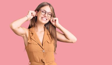 Genç, sarışın, sıradan kıyafetler ve gözlüklerle gülümseyen kulakları parmaklarıyla çeken güzel bir kadın. Komik bir jest. deneme sorunu 