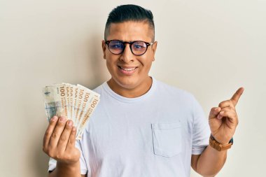 Genç latin adam 100 Danimarka kronu tutuyor gülümsüyor mutlu bir şekilde eli ve parmağı kenarda. 