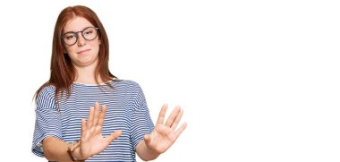 Young read head woman wearing casual clothes and glasses afraid and terrified with fear expression stop gesture with hands, shouting in shock. panic concept. 