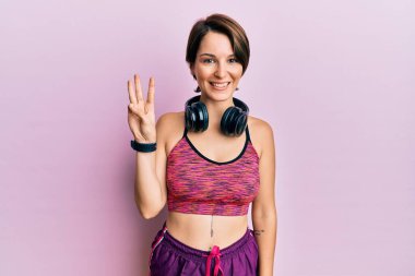 Kısa saçlı, spor giyimli, kulaklık takan, üç numaralı parmaklarıyla kendini beğenmiş ve mutlu gülümseyen esmer kadın.. 