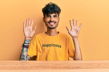 Üzerinde mutluluk mesajı olan İspanyol tişörtü giyen genç adam masanın üzerinde oturuyor ve parmaklarını on numarayı gösterirken kendinden emin ve mutlu bir şekilde gülümsüyor.. 