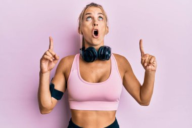 Spor kıyafetleri giyen ve kulaklık takan genç sarışın kadın şaşırmış ve şaşırmış bir şekilde yukarı bakıp parmaklarıyla işaret ediyor ve kollarını kaldırıyordu.. 