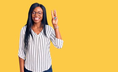 Genç Afro-Amerikalı kadın günlük kıyafetler ve gözlük takıyor. Üç numaralı parmaklarıyla işaret ediyor. Kendinden emin ve mutlu gülümsüyor.. 
