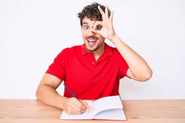 Kıvırcık saçlı genç beyaz adam masaya oturmuş defter yazıyor. Gülümsüyor ve gülümsüyor. Tamam. 