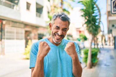 Orta yaşlı, kır saçlı, günlük kıyafetler giyen, mutlu ve heyecanlı bir şekilde kolları havada, gülümseyen ve başarı çığlıkları atan bir adam. kutlama konsepti. 