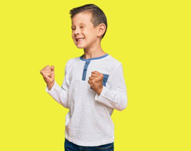 Sevimli beyaz çocuk, gündelik kıyafetler giyip çok mutlu ve heyecanlı bir şekilde kollarını kaldırıp, gülümseyerek ve başarı için bağırarak kazanan jestler yapıyor. kutlama konsepti. 