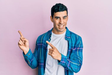 Yakışıklı, sıradan kıyafetler giyen, gülümseyen ve kameraya bakan, iki elini ve parmağını yana doğru gösteren İspanyol bir adam.. 