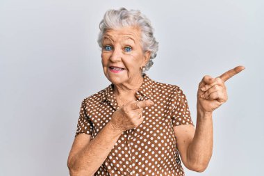 Yaşlı, gri saçlı, günlük kıyafetler giyen, gülümseyen ve kameraya bakan iki eliyle ve parmaklarıyla yüzünü gösteren bir kadın.. 