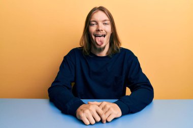 Uzun saçlı, sıradan kıyafetler giyen yakışıklı beyaz adam masada oturmuş komik bir ifadeyle dilini dışarı çıkarıyor. duygu kavramı. 