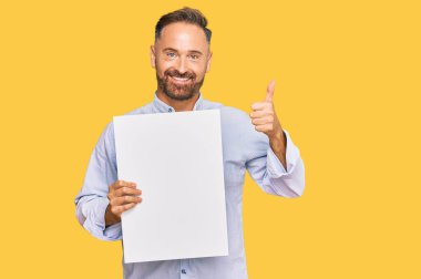 Yakışıklı, orta yaşlı, elinde boş bir afiş tutan mutlu ve olumlu gülümseyen, başparmağıyla mükemmel ve onay işareti yapan bir adam. 