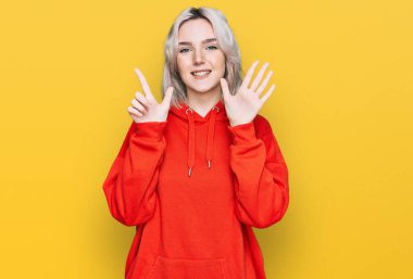 Genç sarışın kız gündelik kıyafetler giyer ve yedinci parmaklarını göstererek gülümserken kendinden emin ve mutluydu.. 