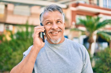 Orta yaşlı, ispanyol, kır saçlı, gülümseyen, mutlu, parkta akıllı telefondan konuşan bir adam..