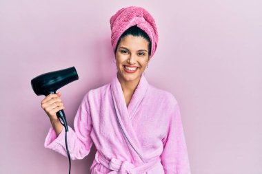 Duşta bornoz giyen genç İspanyol kadın kurutucu kullanıyor. Pozitif görünüyor ve gülümsüyor. Kendine güvenen bir gülümsemeyle dişlerini gösteriyor. 