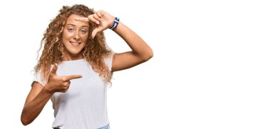 Güzel beyaz tişörtlü beyaz kız gülümseyerek elleri ve parmakları gülen yüzlü bir çerçeve yapıyor. yaratıcılık ve fotoğrafçılık kavramı. 