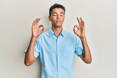 Genç, yakışıklı, Afro-Amerikalı, günlük kıyafetler giymiş rahat ve gözleri kapalı gülümseyen parmaklarıyla meditasyon hareketi yapan bir adam. yoga konsepti. 