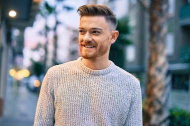 Genç beyaz adam gülümseyerek şehirde mutlu mesut duruyor..