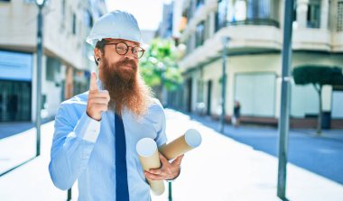 Uzun sakallı, kasketli, mutlu gülümseyen genç bir mimar. Yüzünde tebessüm, elinde planlar ve parmağınla şehrin caddesindeki kamerayı gösteriyorsun..