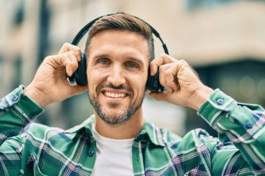 Genç beyaz adam şehirde kulaklık takarak mutlu bir şekilde gülümsüyor..