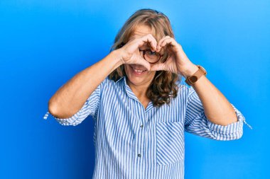 Orta yaşlı, sarışın, günlük kıyafetler ve gözlük takan, kalp şeklinde el ve parmakları olan, tabelanın içinden bakıp gülümseyen bir kadın. 