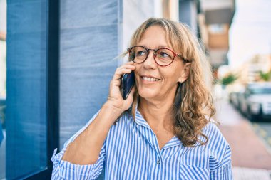 Orta yaşlı, beyaz, gülümseyen bir kadın. Şehirdeki akıllı telefondan mutlu bir şekilde konuşuyor..