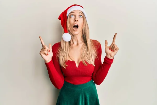 穿着圣诞服装和帽子的美丽的高加索女人惊奇地抬起头 用手指和胳膊指指着 — 图库照片