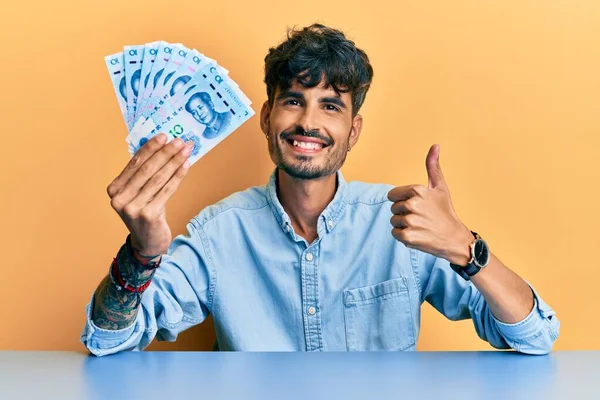 Joven Hispano Sosteniendo Billetes Chinos Yuan Sentado Mesa Sonriendo Feliz —  Fotos de Stock